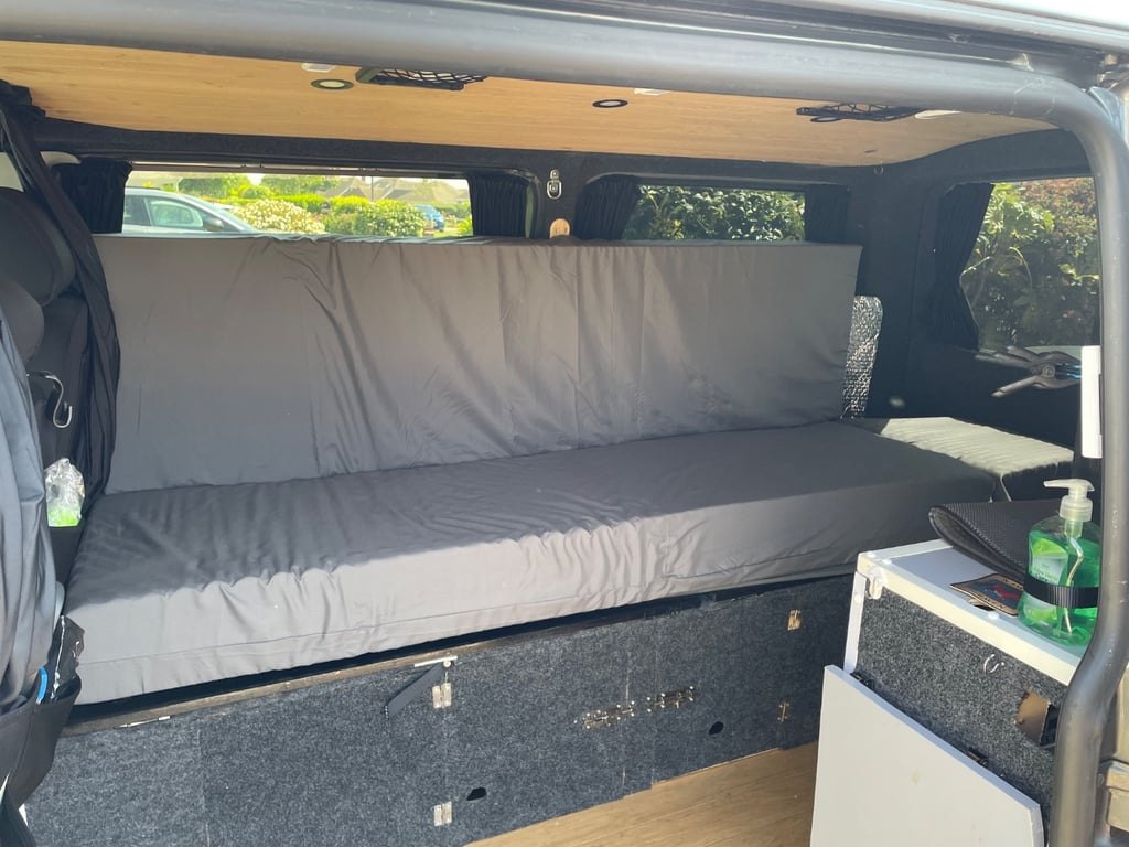The campervan's bed made up with a mattress and cushions along the side of the van, next to a window