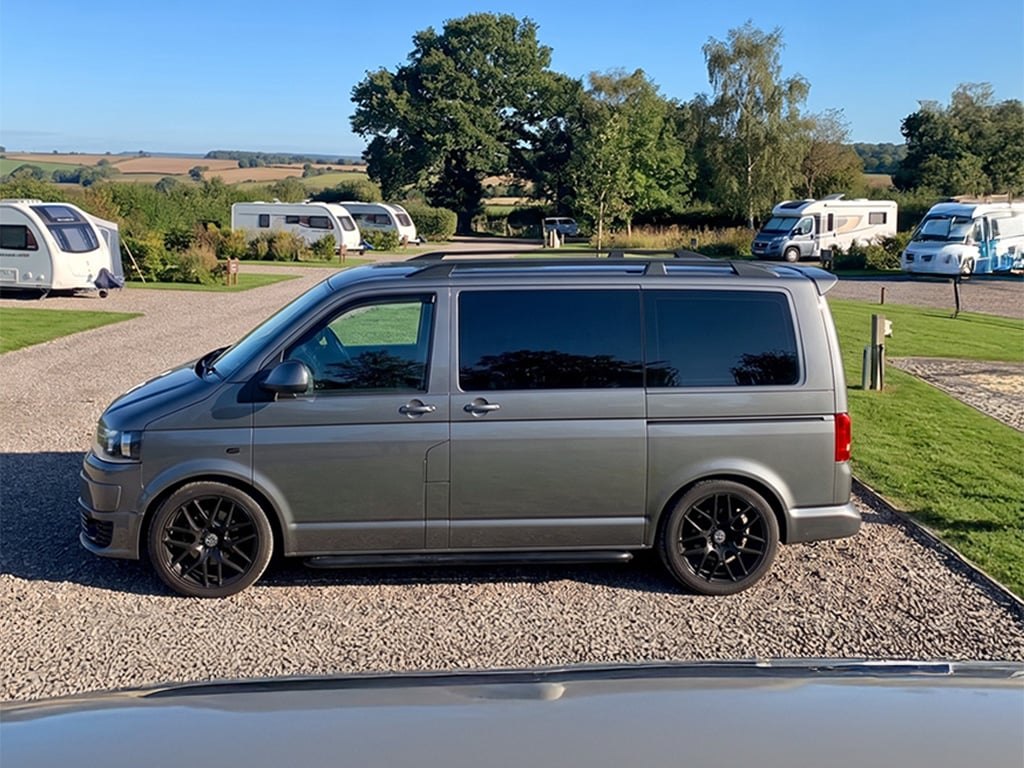 The grey VW Transporter campervan parked side-on at a campsite pitch with white caravans and rolling countryside behind