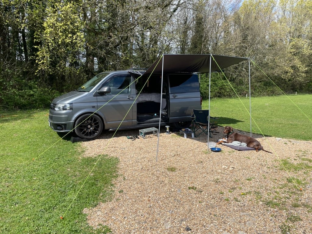 The campervan parked on grass with a sun canopy extended from the side, a dog lying in the shade underneath and camping chairs set out