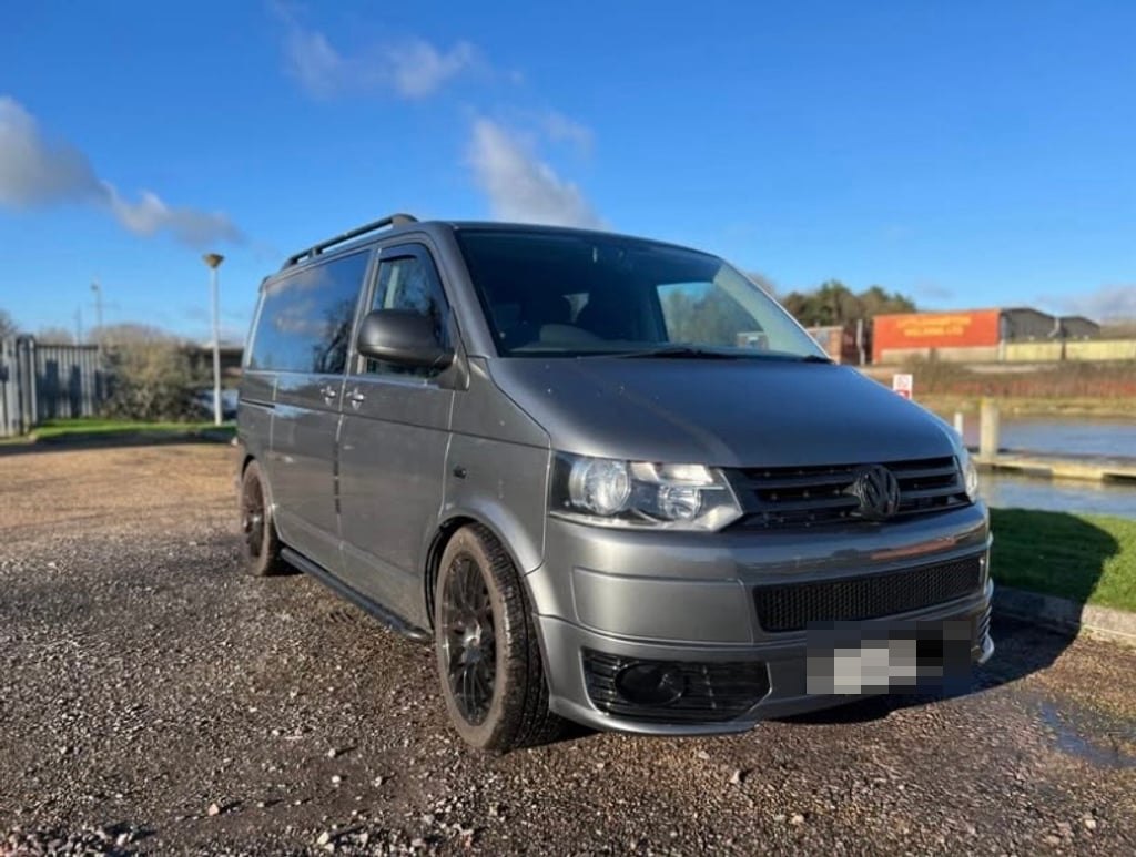 Front three-quarter view of the grey VW Transporter T5.1 campervan on gravel beside water, with the VW badge and black grille visible (number plate digitally obscured)