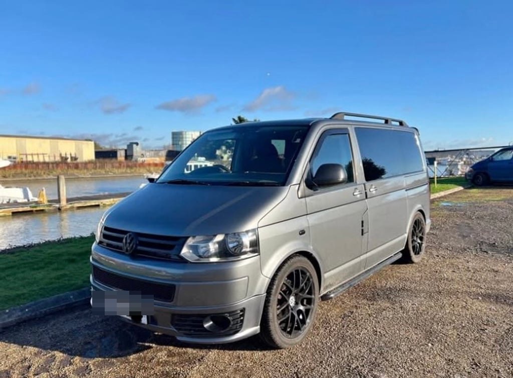 A grey 2010 Volkswagen Transporter T5.1 campervan on black alloy wheels, parked on gravel beside a harbour under a clear blue sky (number plate digitally obscured)