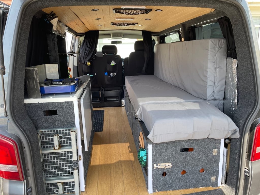 The rear interior of the campervan seen through the open tailgate, showing the bed platform, storage boxes and the cab beyond