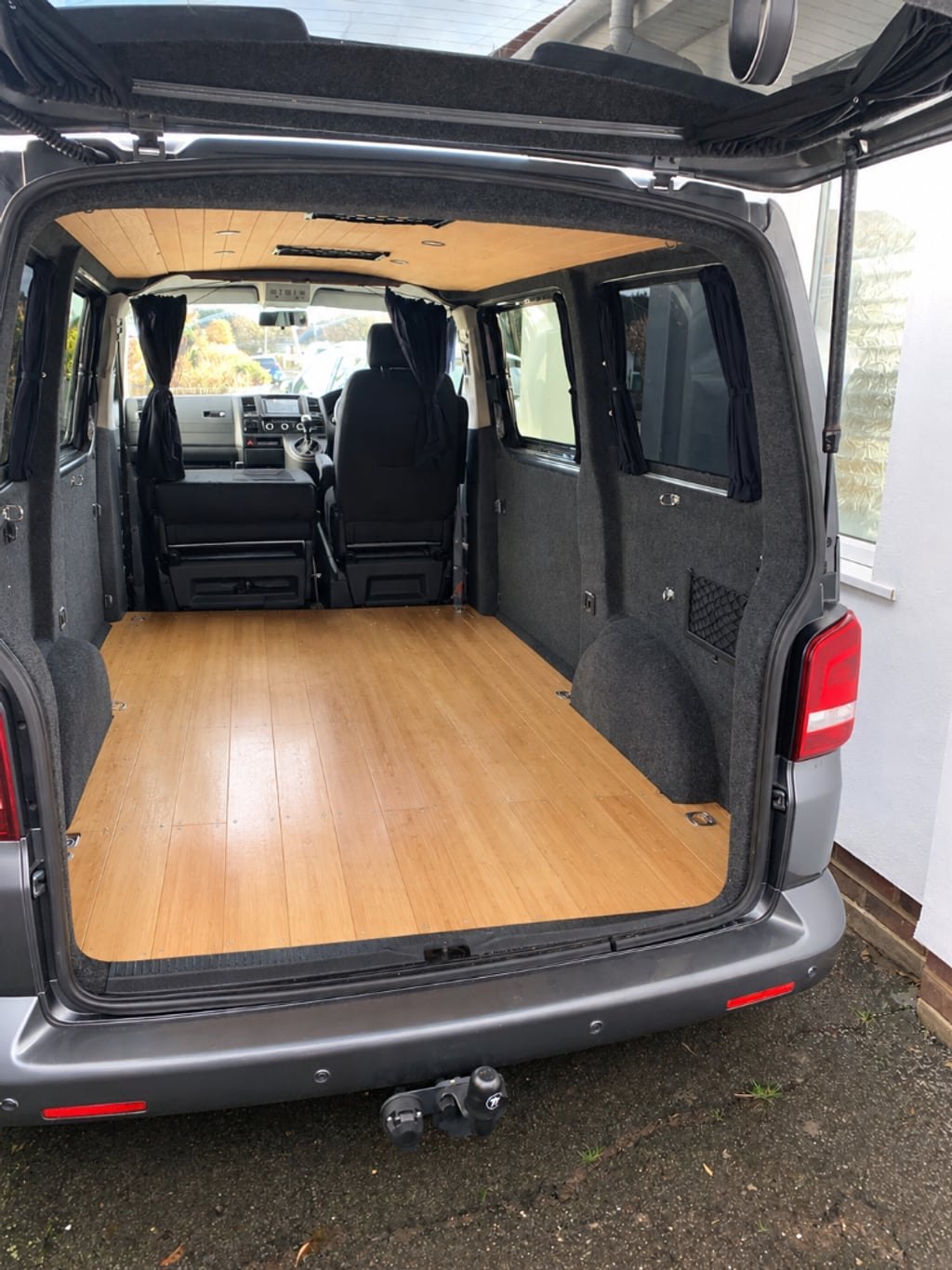 The campervan with its tailgate open and rear doors wide, showing the bamboo-effect wooden floor, curtained windows and cab seats beyond