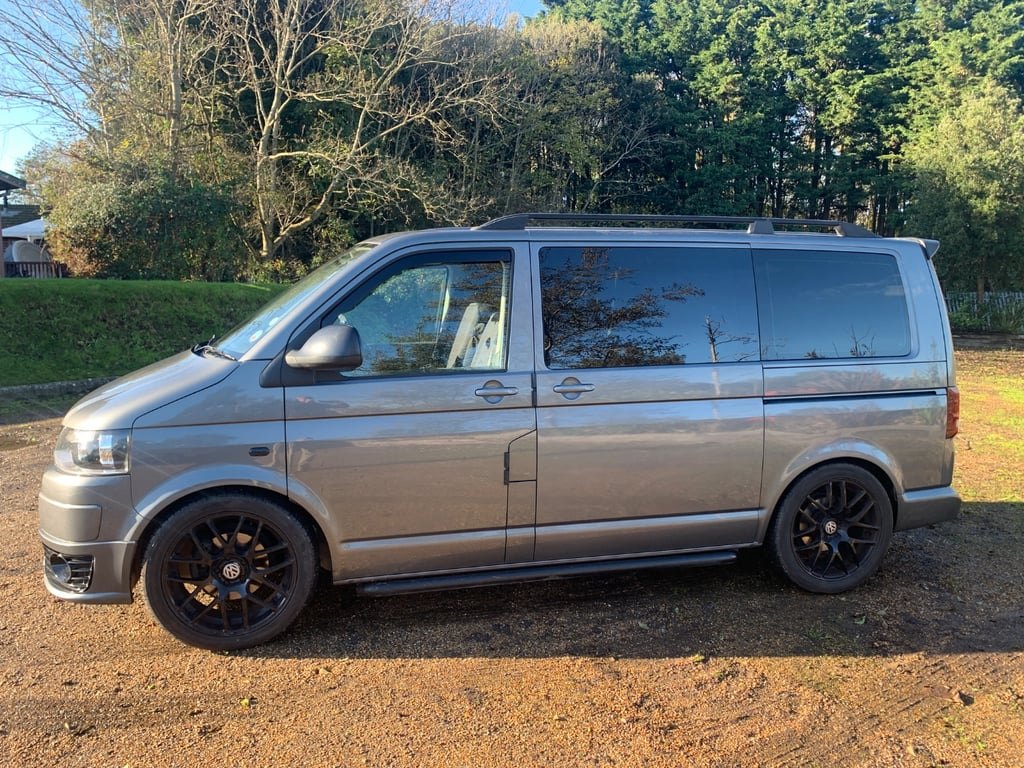 Side profile of the grey VW Transporter T5.1 campervan with black alloy wheels and side bars, parked on gravel in front of woodland