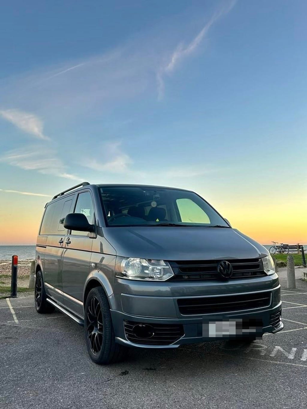 The VW Transporter T5.1 campervan parked facing the sea at sunset, with an orange and blue sky behind it (number plate digitally obscured)